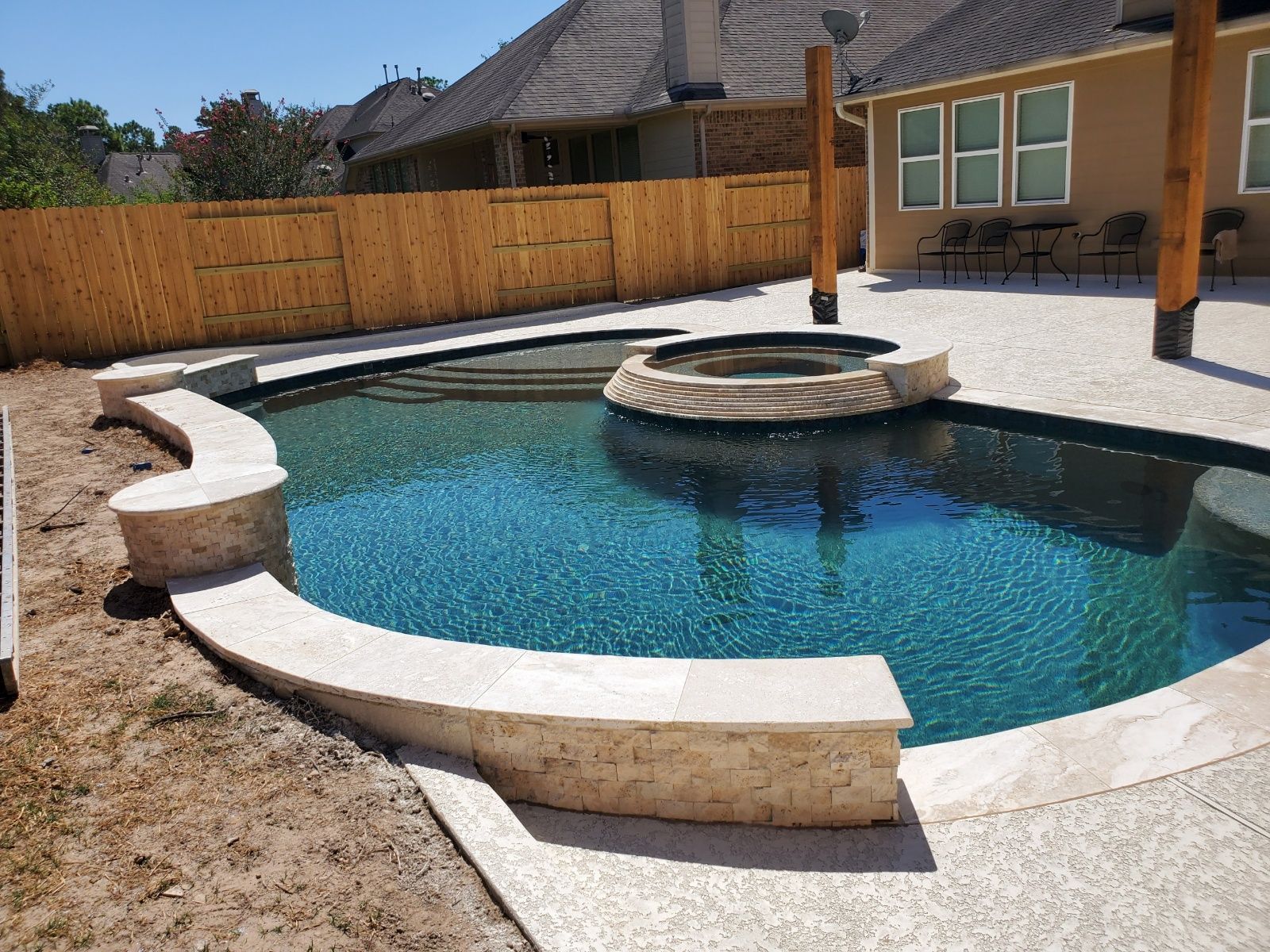 Backyard swimming pool with curved stone deck and a raised circular spa, fenced yard and house in background