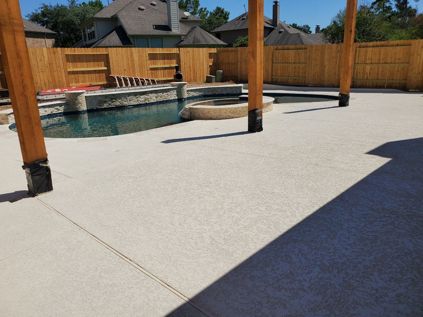 Covered patio with a pool, wooden fence, and concrete deck in bright sunlight
