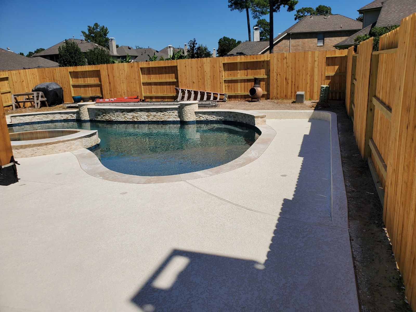 Backyard pool with light stone deck, wooden fence, and bright blue sky