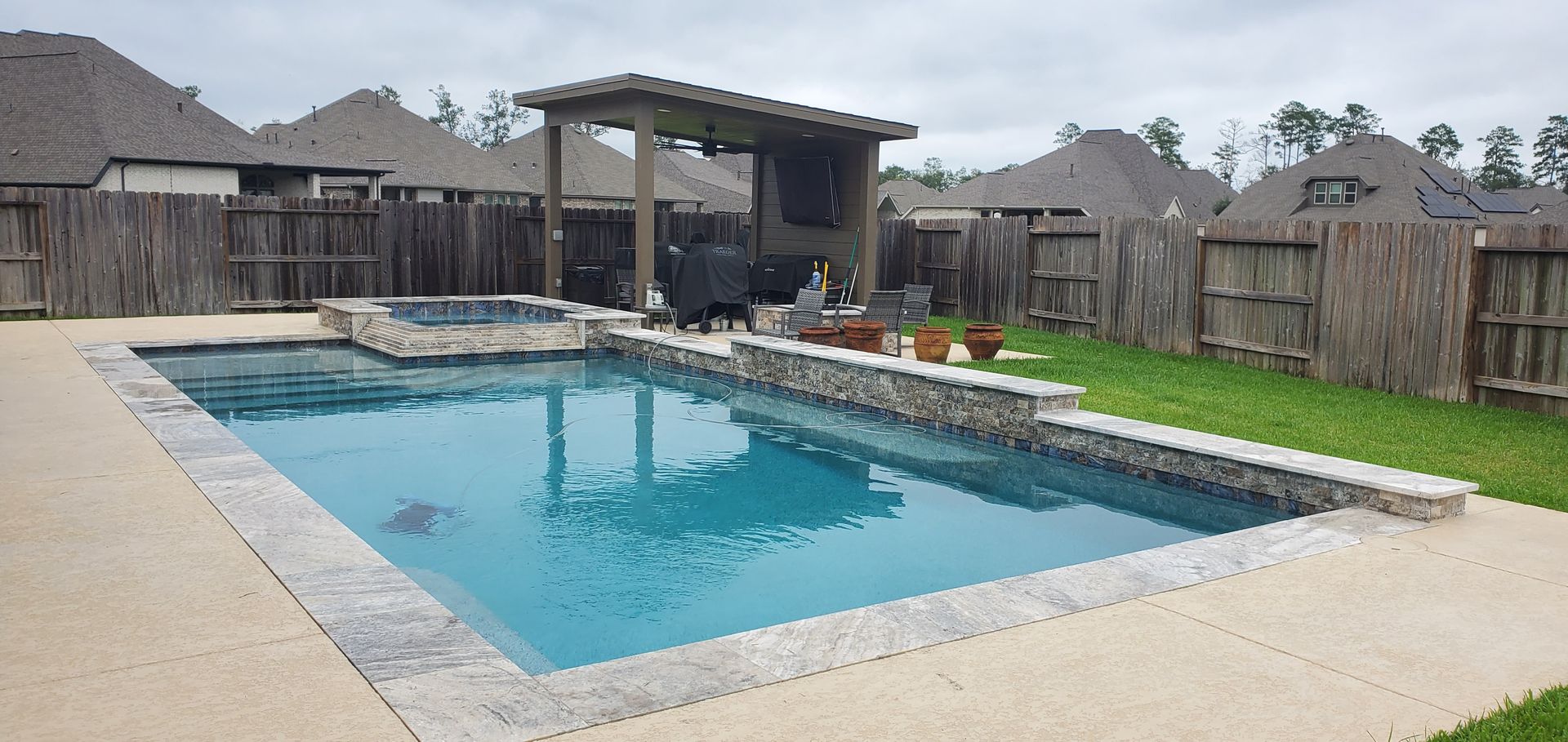 Backyard swimming pool with covered patio, stone deck, and fenced lawn under overcast sky