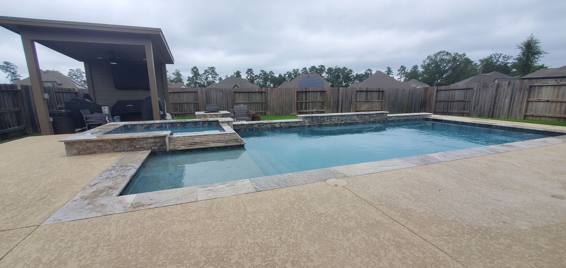 Backyard swimming pool beside a covered patio and fenced yard under a cloudy sky