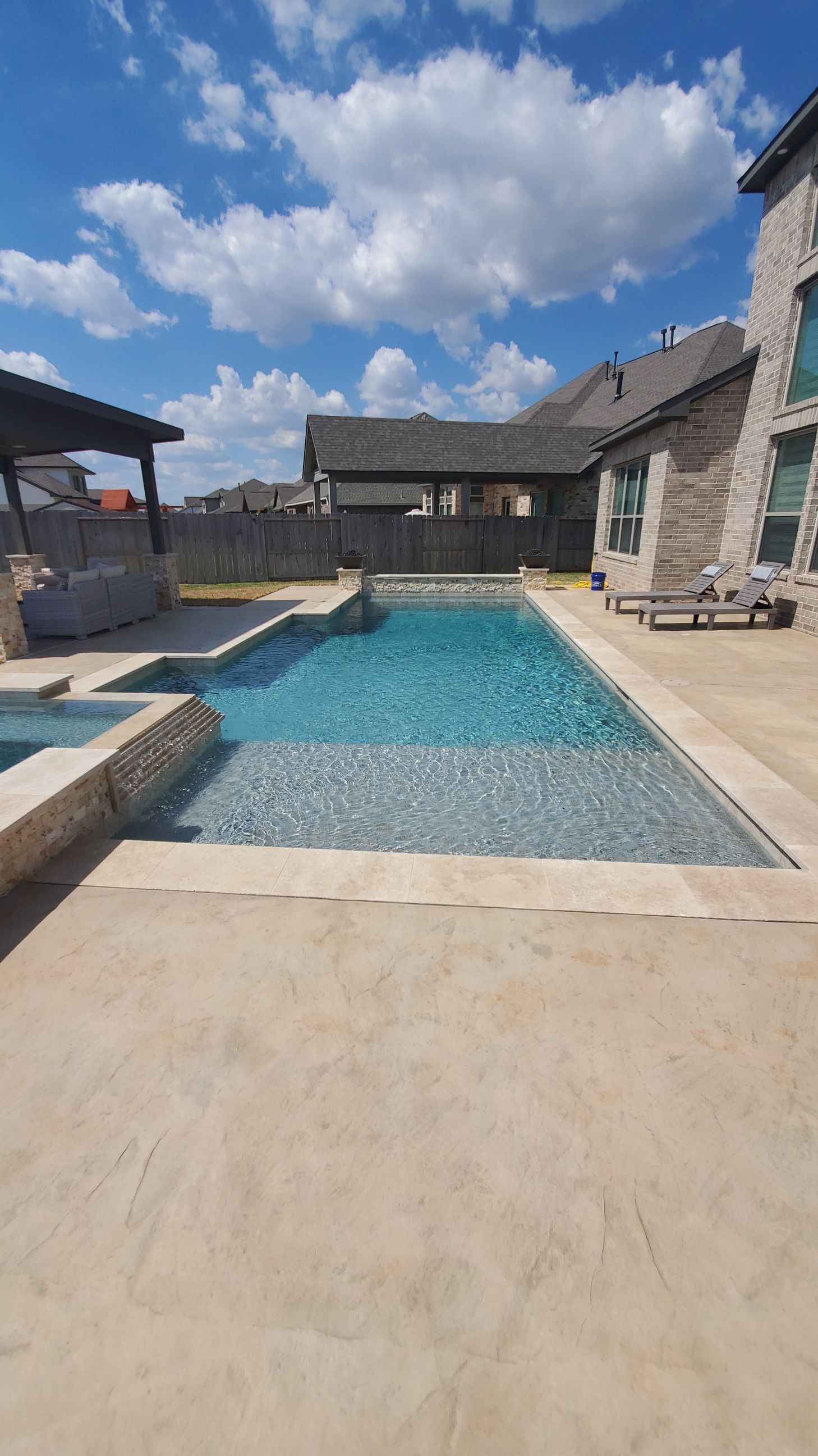 Backyard pool with stone patio, blue water, and stone houses under a partly cloudy sky