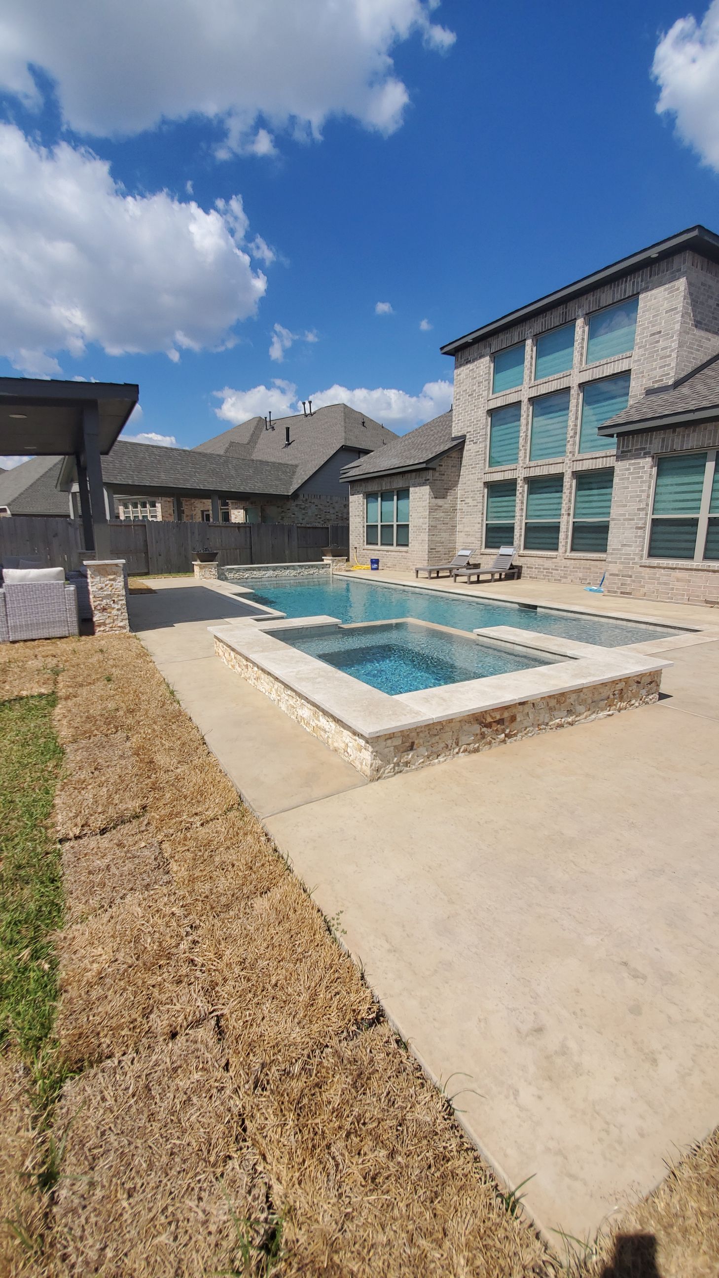 Backyard patio with a small rectangular pool, stone house, and blue sky with clouds.