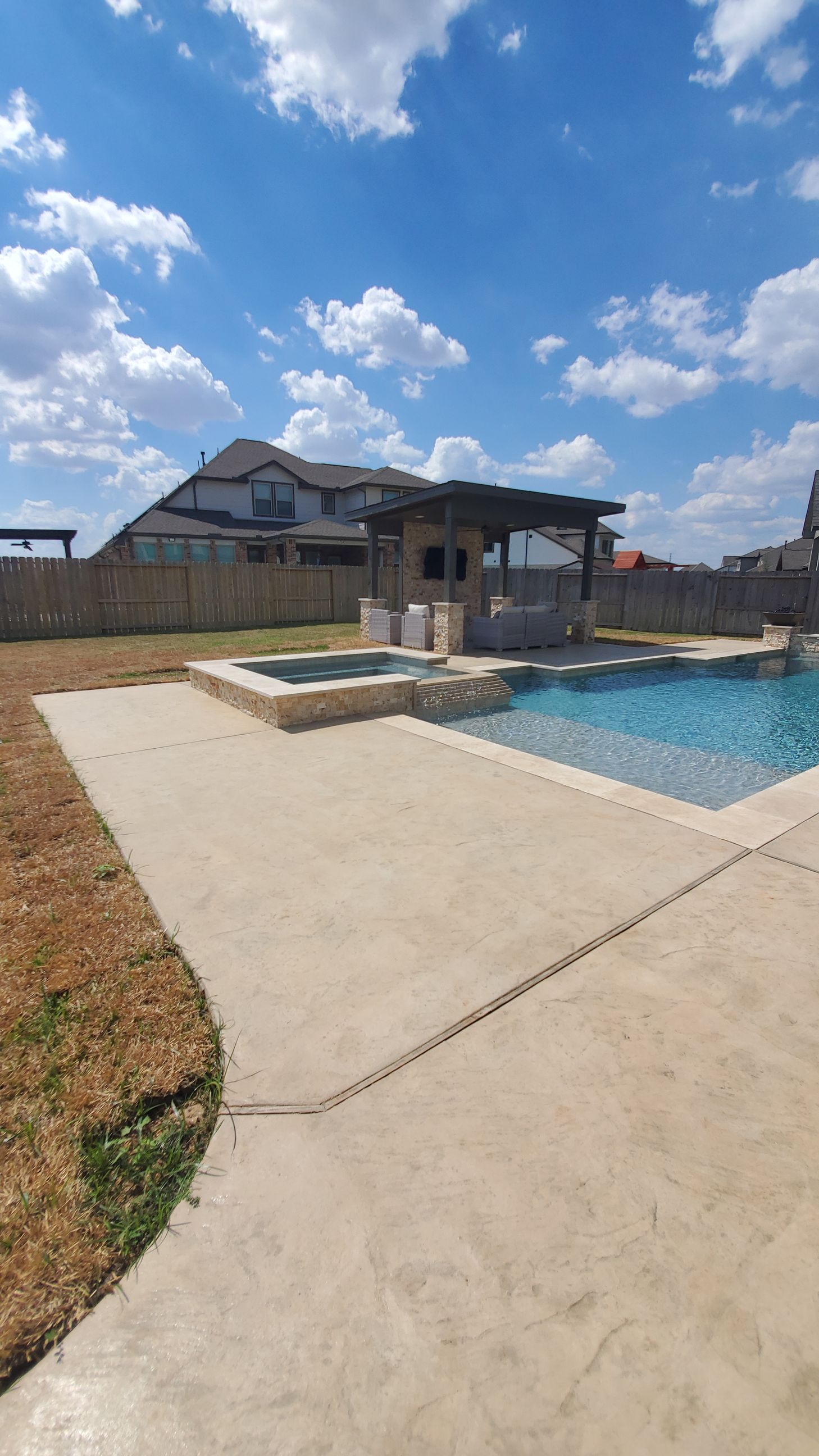 Backyard pool with concrete patio, fence, and a house under a bright blue sky with clouds