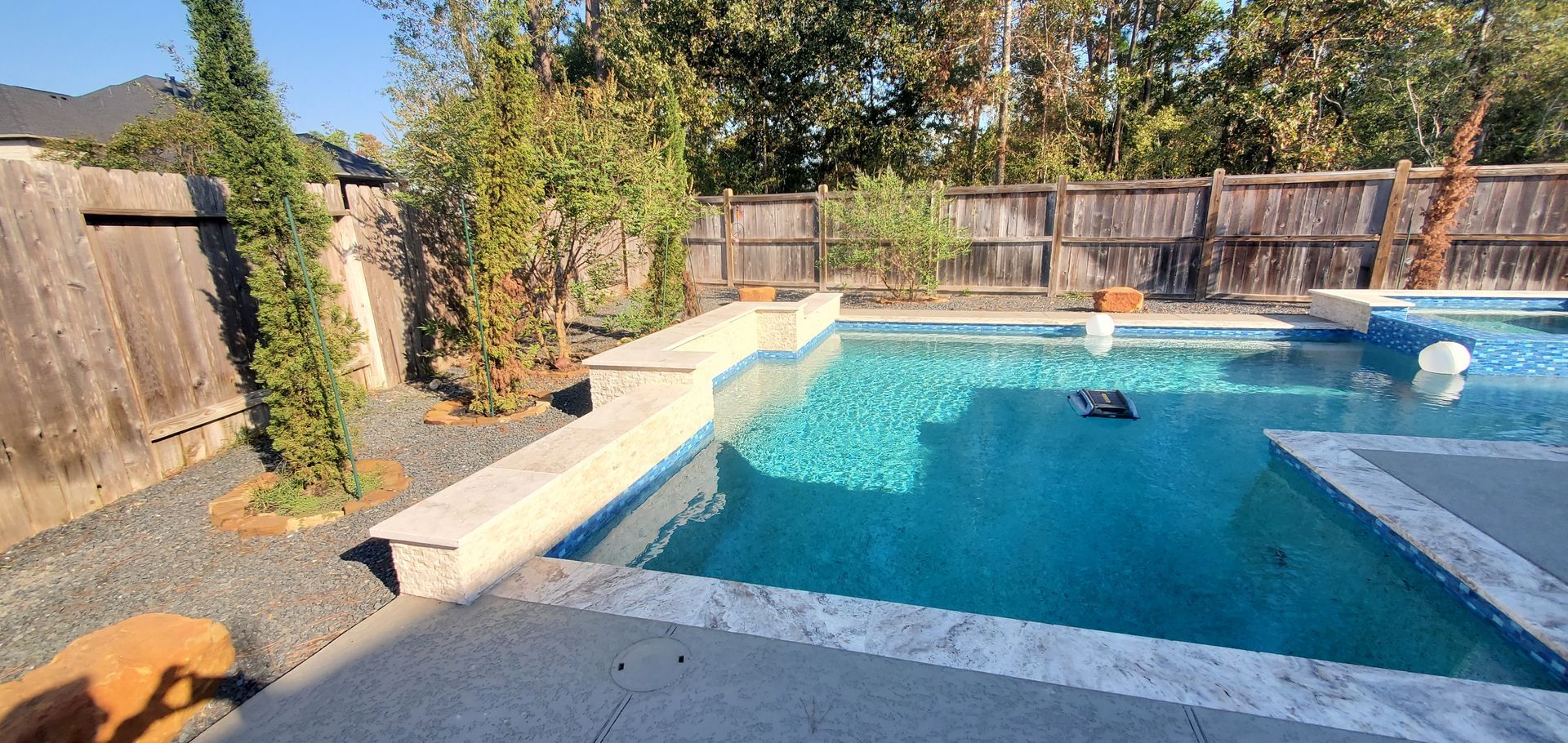 Backyard swimming pool with blue water, stone coping, wooden fence, and trees in sunlight