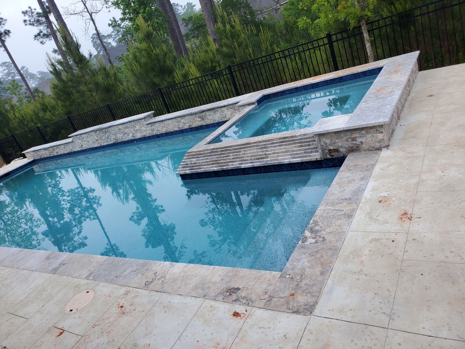 Backyard swimming pool with a raised spa and stone deck, surrounded by greenery and a black fence