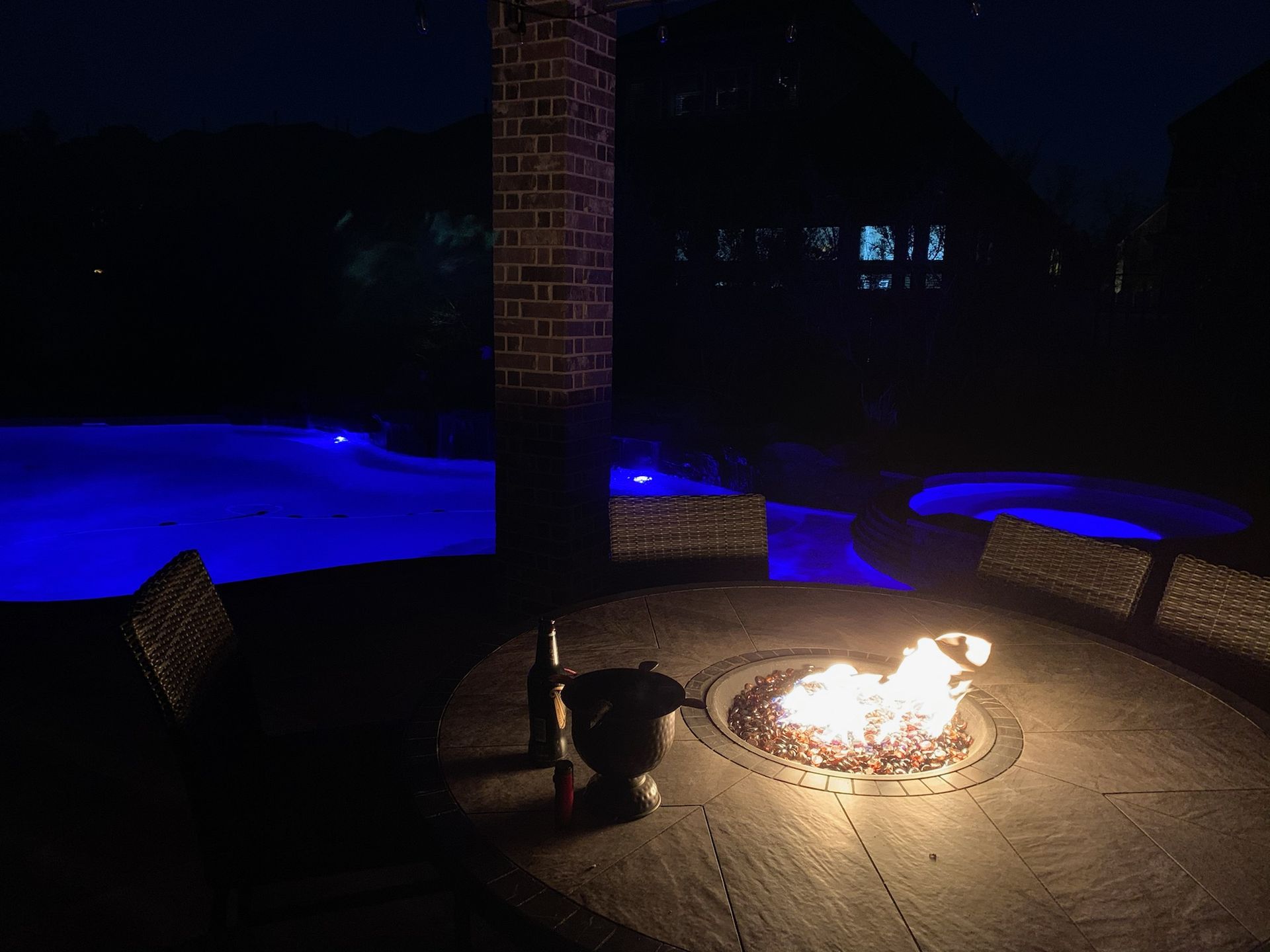 Night patio with a fire pit, chairs, and a lit blue swimming pool in the background