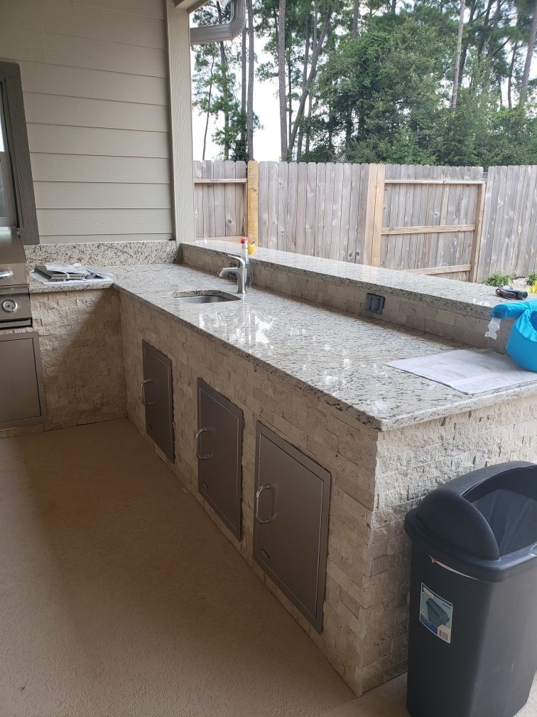 Outdoor stone countertop with built-in sink and cabinets on a covered patio, with a fence and trees behind it