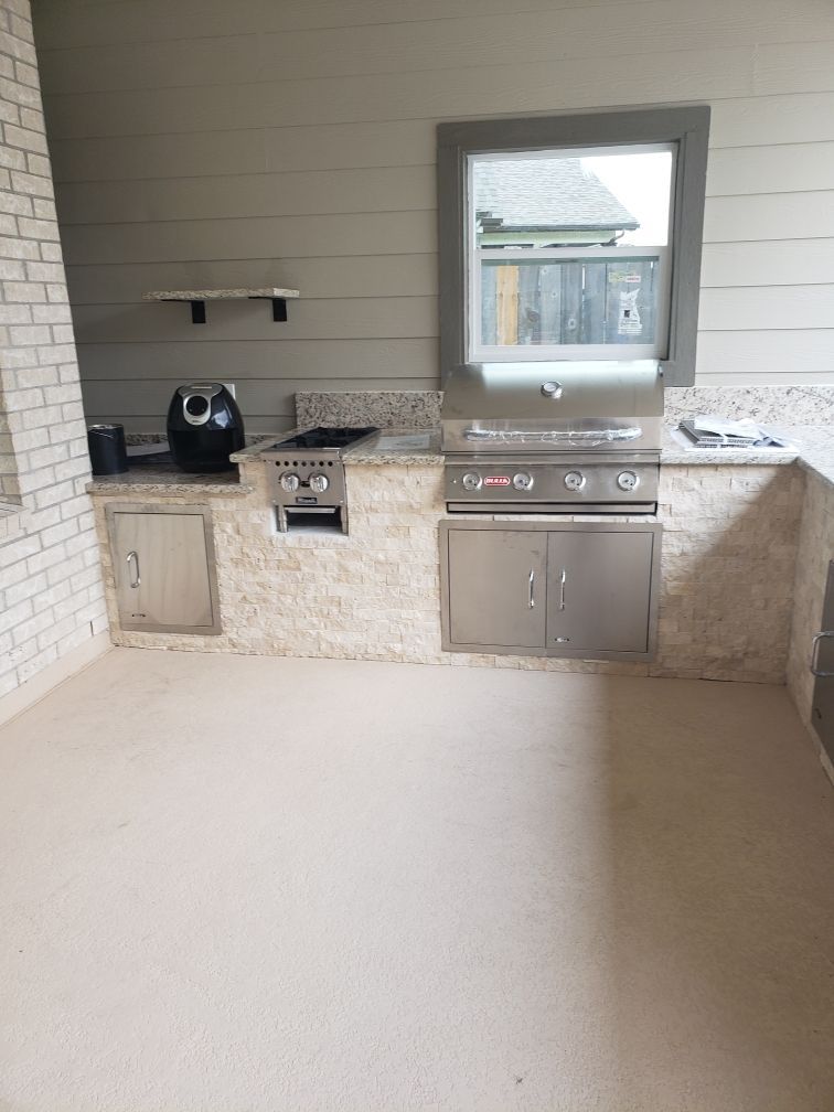 Modern outdoor kitchen with grill, sink, stone counters, and a window overlooking a yard.