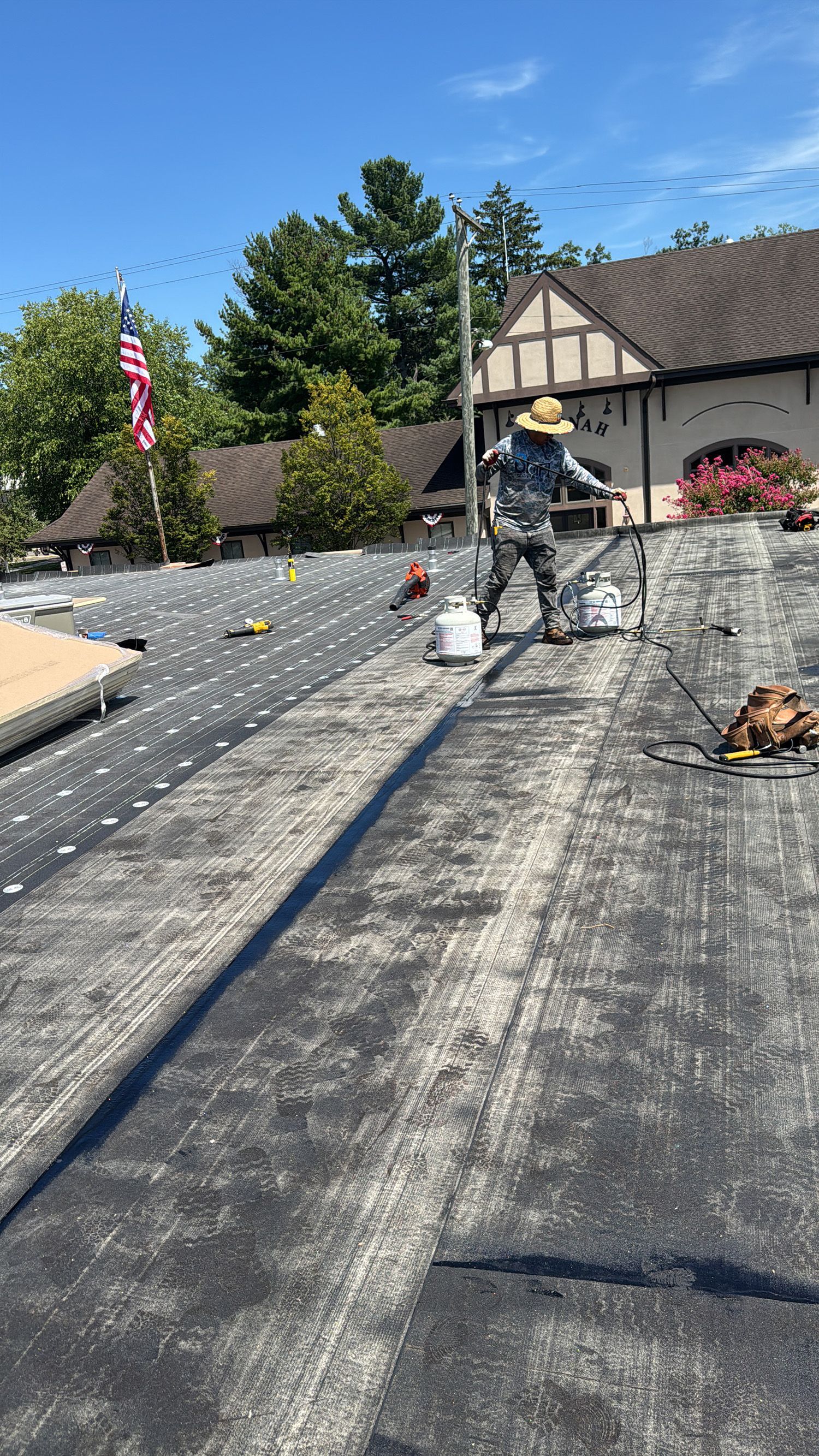 Roofer on a dark-colored, weathered roof, working in bright sunlight with tools and a flag in the background.