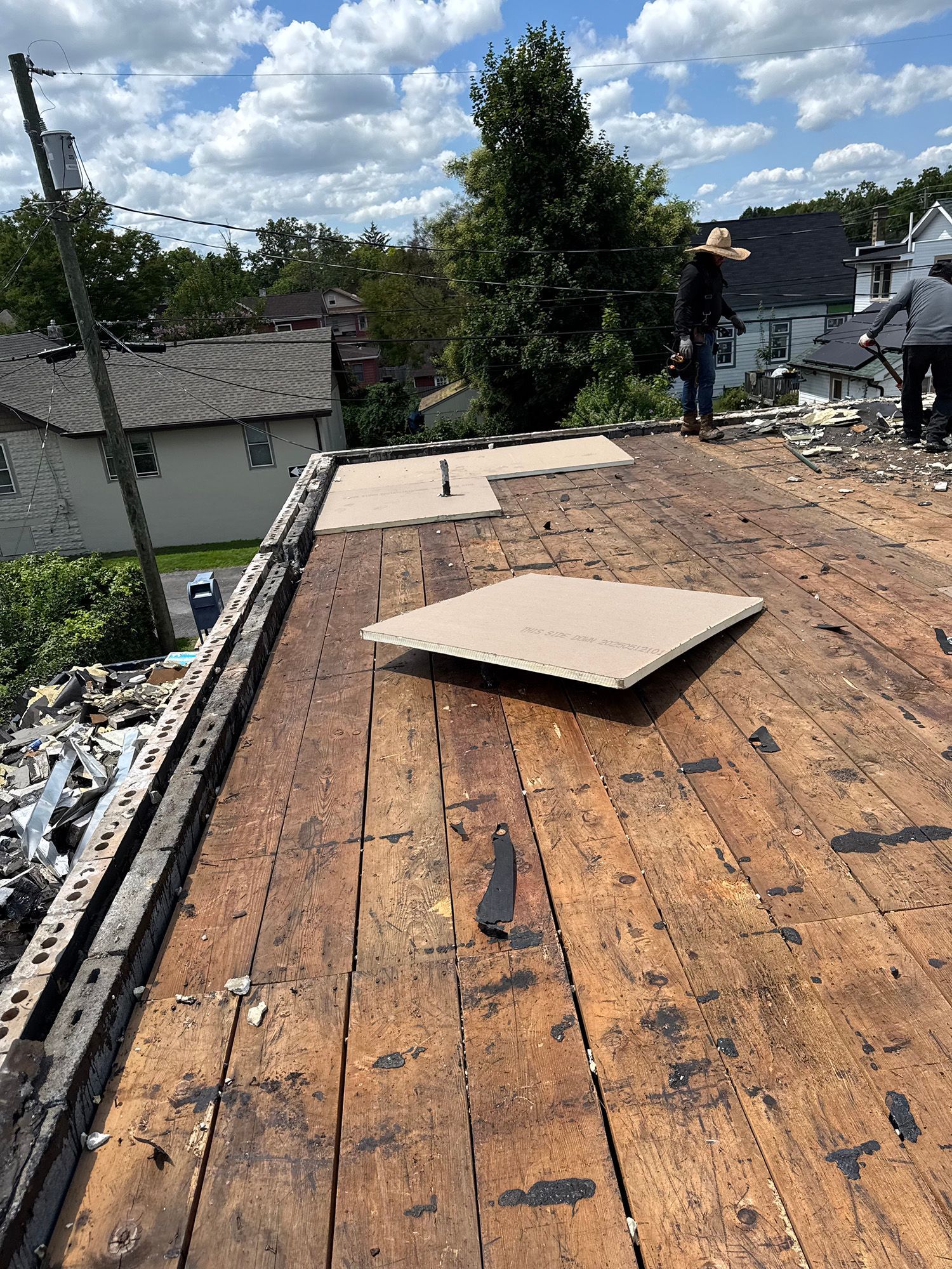 Workers on a roof removing old shingles. Brown wood deck, blue sky, trees in background.