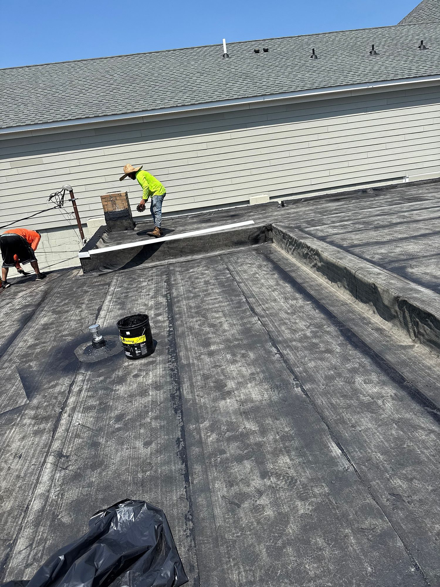 Roofers working on a flat roof, applying sealant. One in a yellow vest, on a sunny day.