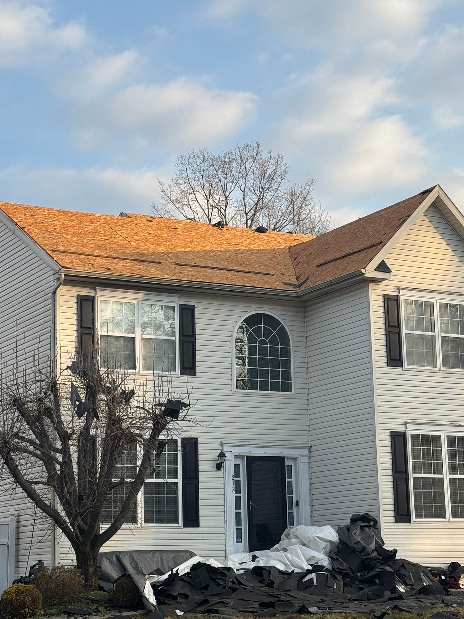 Two-story house with new brown roof; pile of roofing material on the ground. Cloudy sky.
