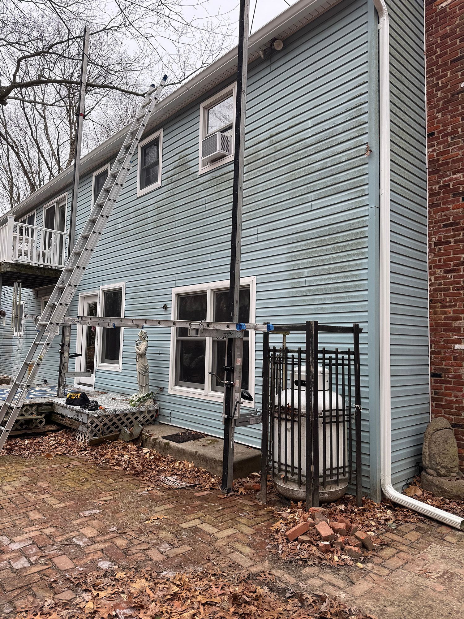 Two-story blue house with a ladder, scaffolding, and propane tank. Brick pathway and fallen leaves on the ground.
