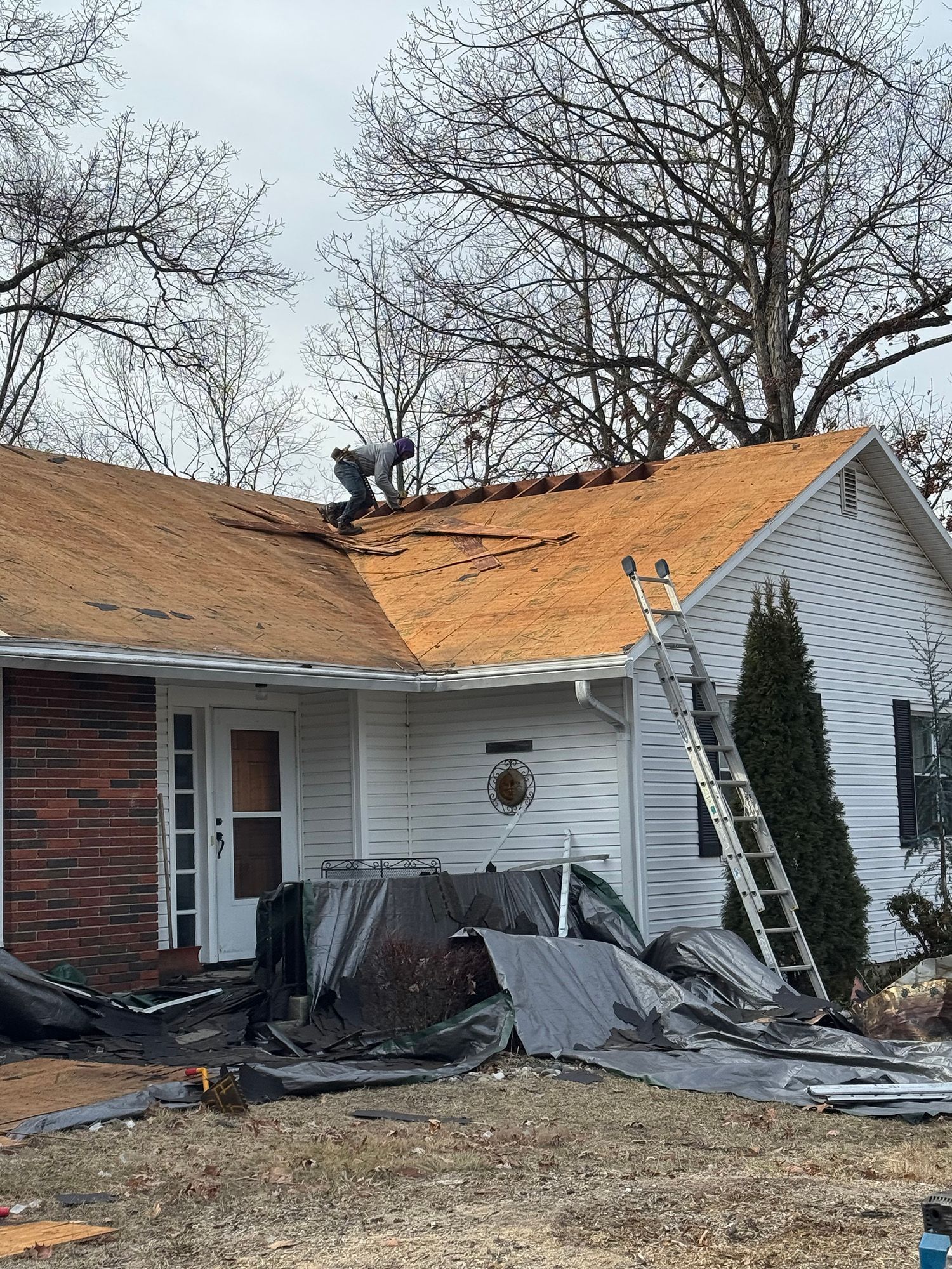 A worker on a roof replacing shingles on a white house with a brick accent.