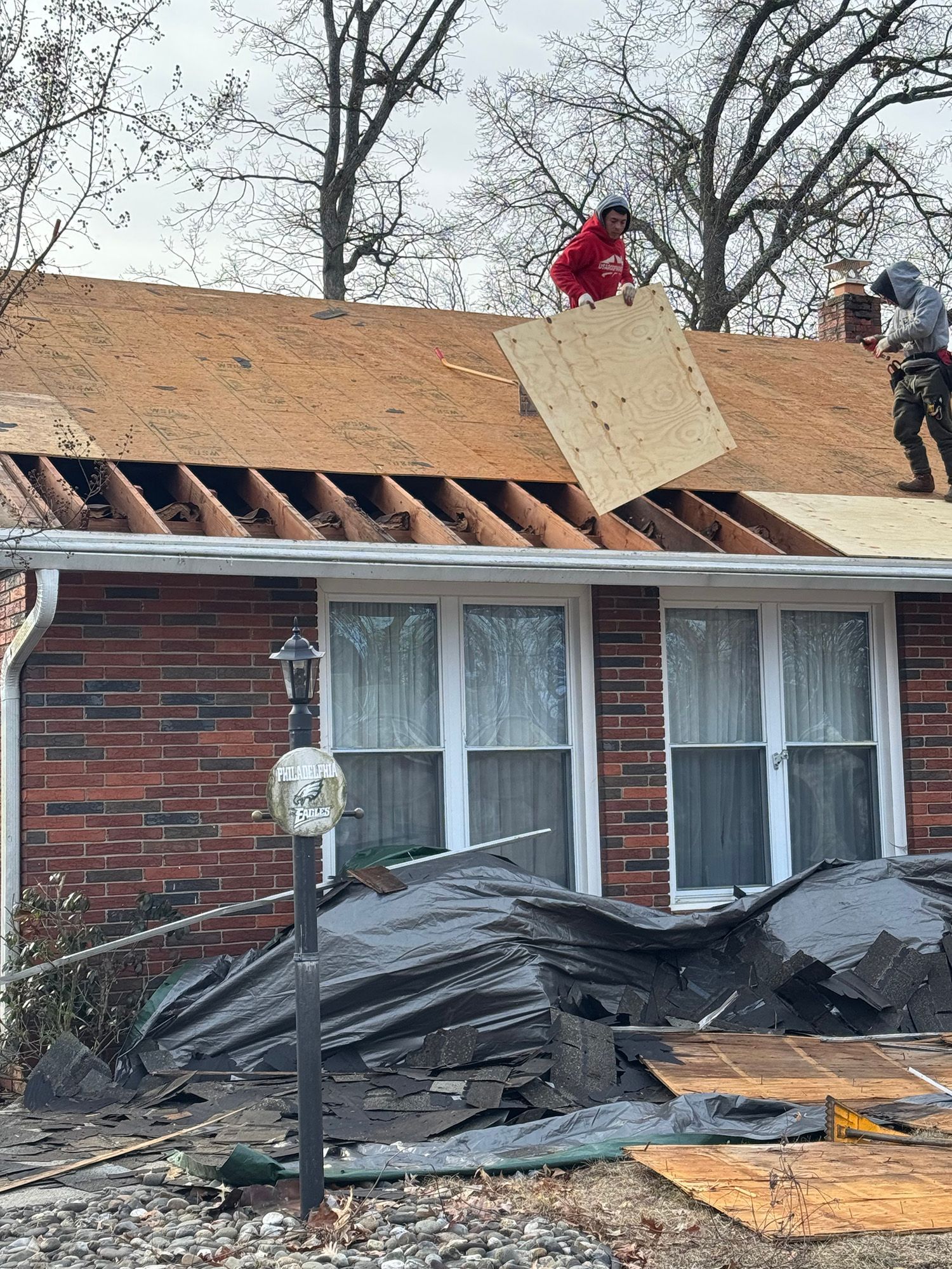 Roofers replacing a roof; one holds plywood while another works. Debris on ground, red brick house.