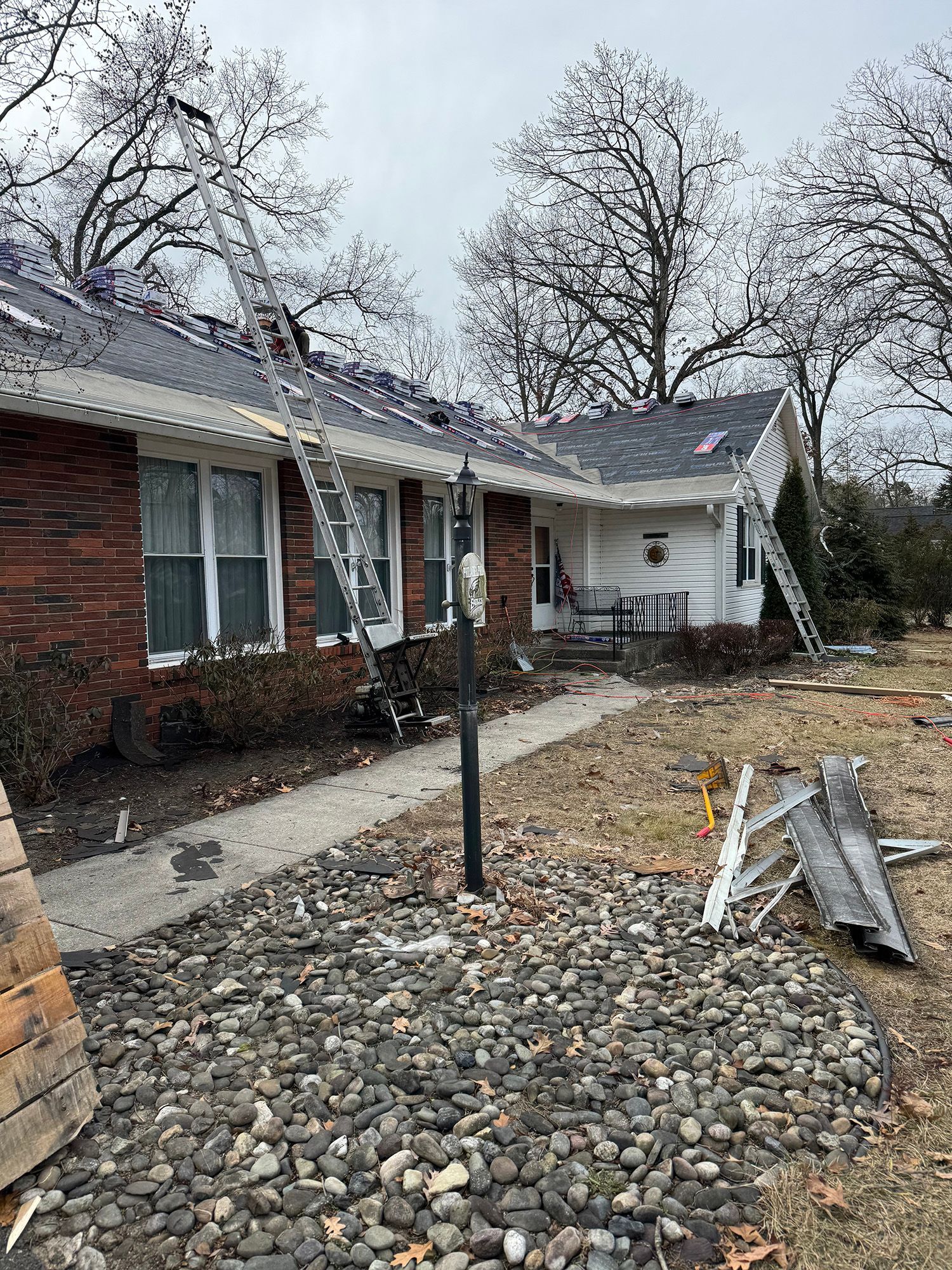 House with ladders on the roof; construction work in progress. Brick facade, cloudy sky, debris in yard.