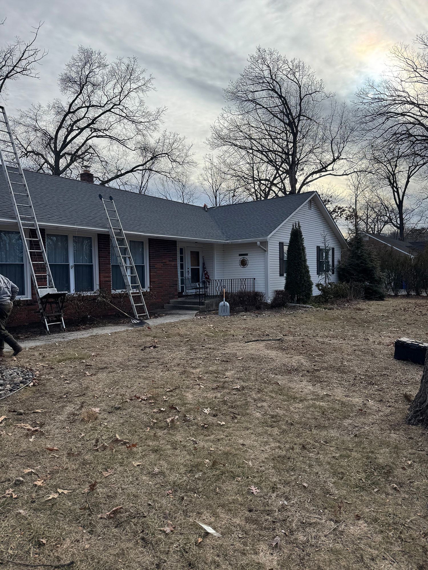 A house with a newly shingled roof, two ladders, and bare trees on a cloudy day.