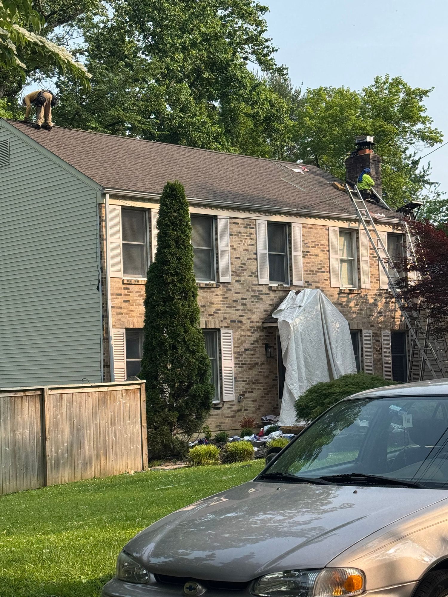 Workers on a roof, ladder propped up. Brick house with white shutters, covered entryway.