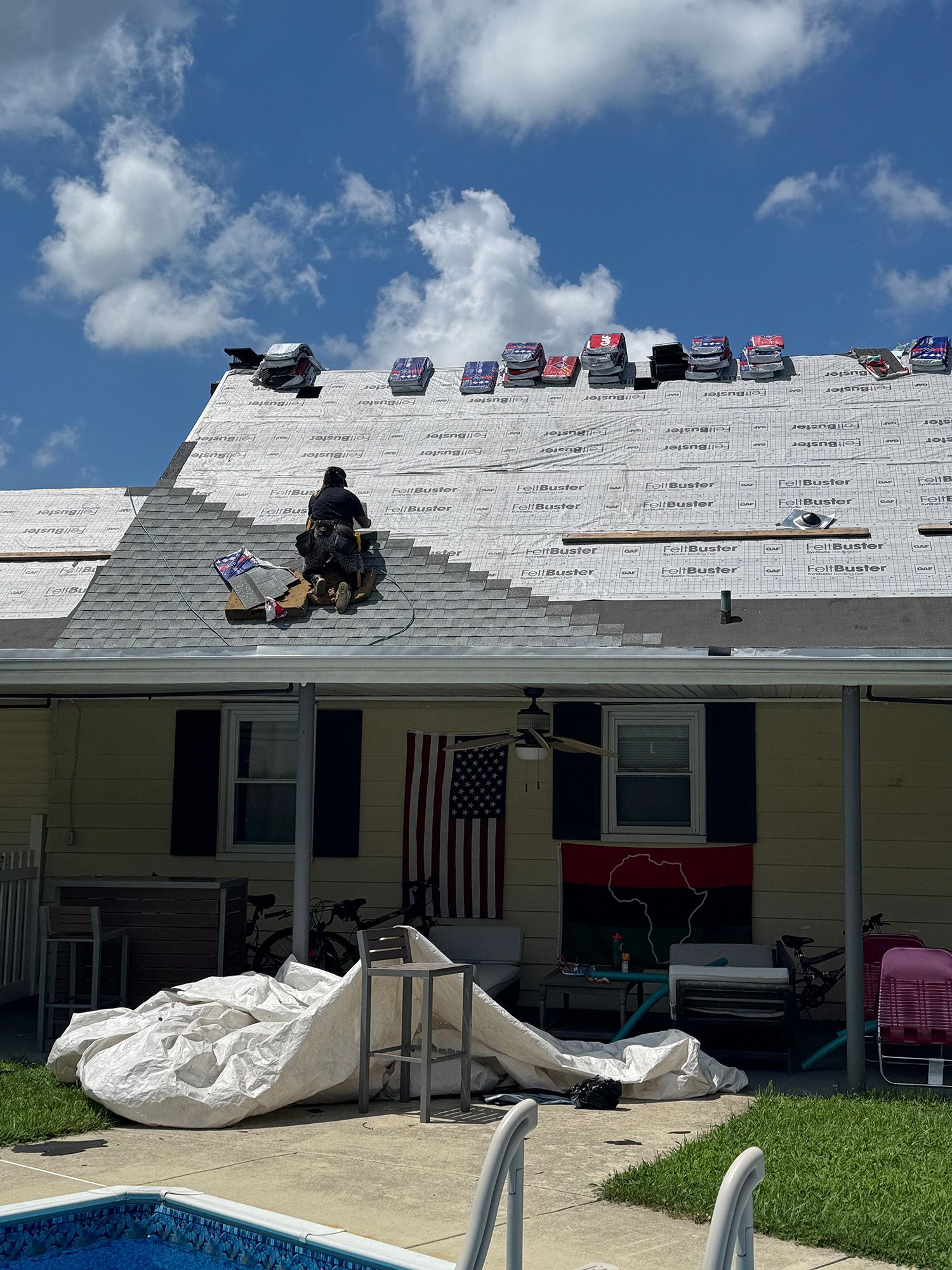 Man on roof replacing shingles on a sunny day with an American flag visible.