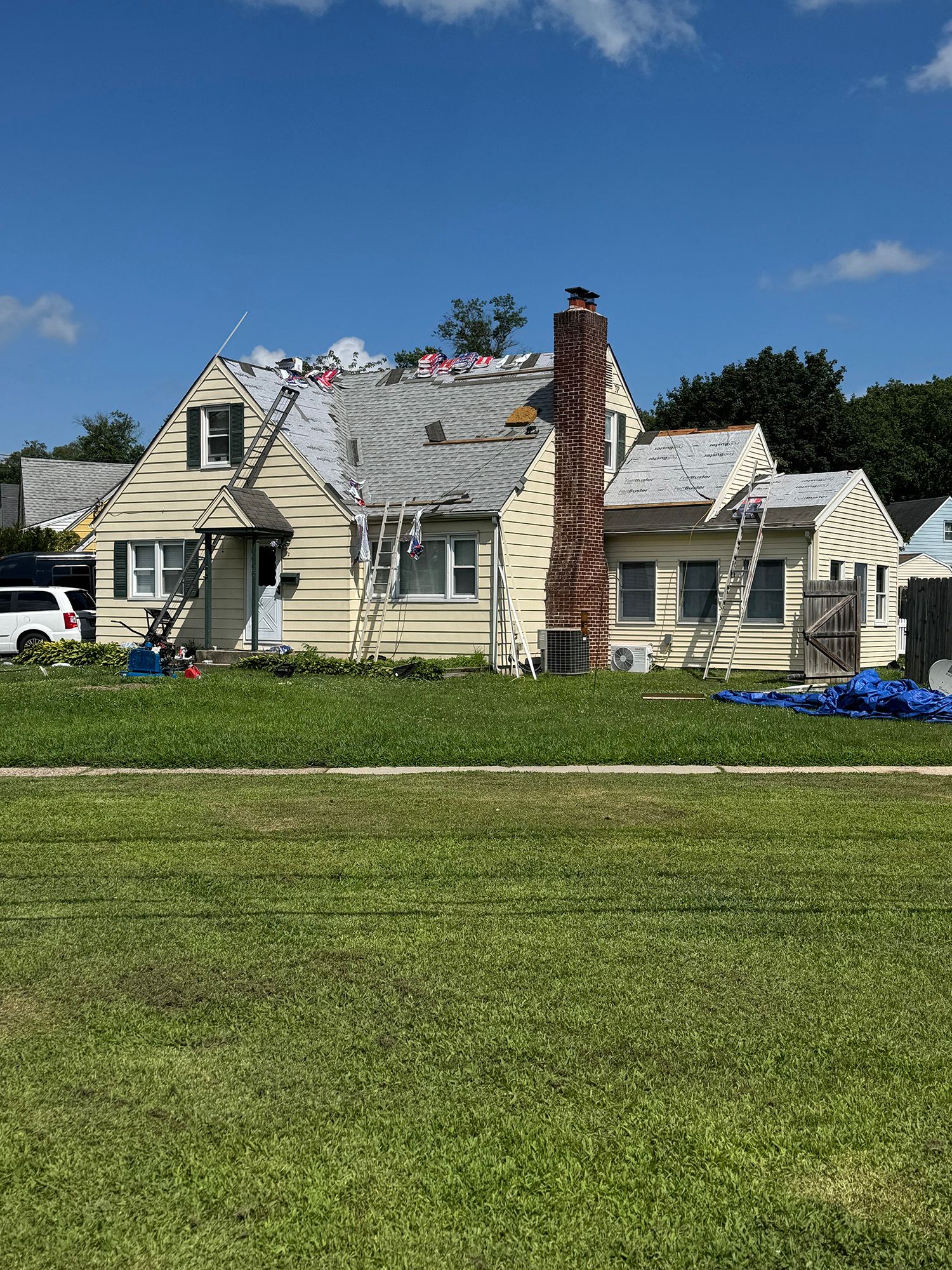 House with partially removed roof; ladders, construction materials, and chimney visible. Sunny day, green lawn.
