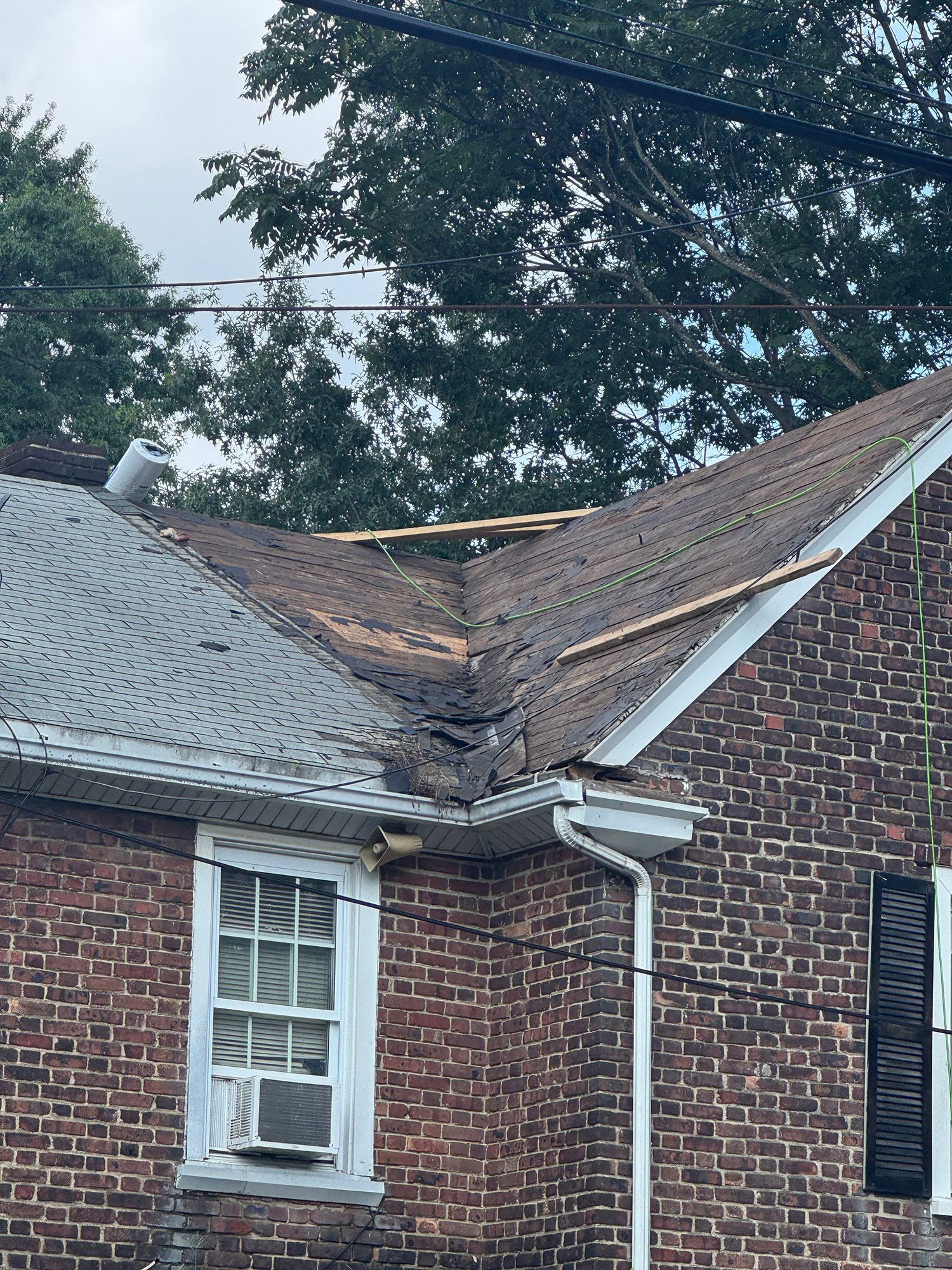 Brick house with damaged roof and exposed wood, window with AC, and dark shutters.