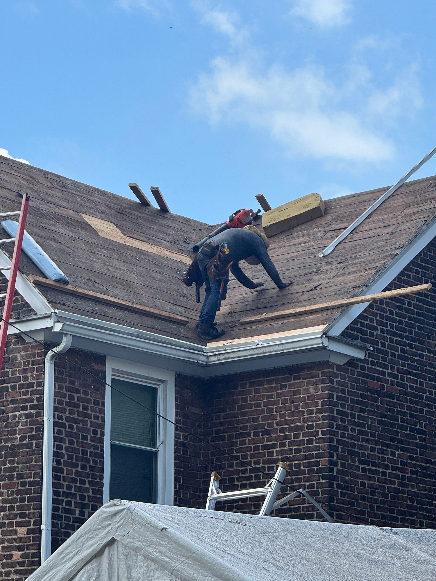 Two workers on a brown shingled roof, near a brick building. One cuts with a saw, the other kneels. Bright sky.