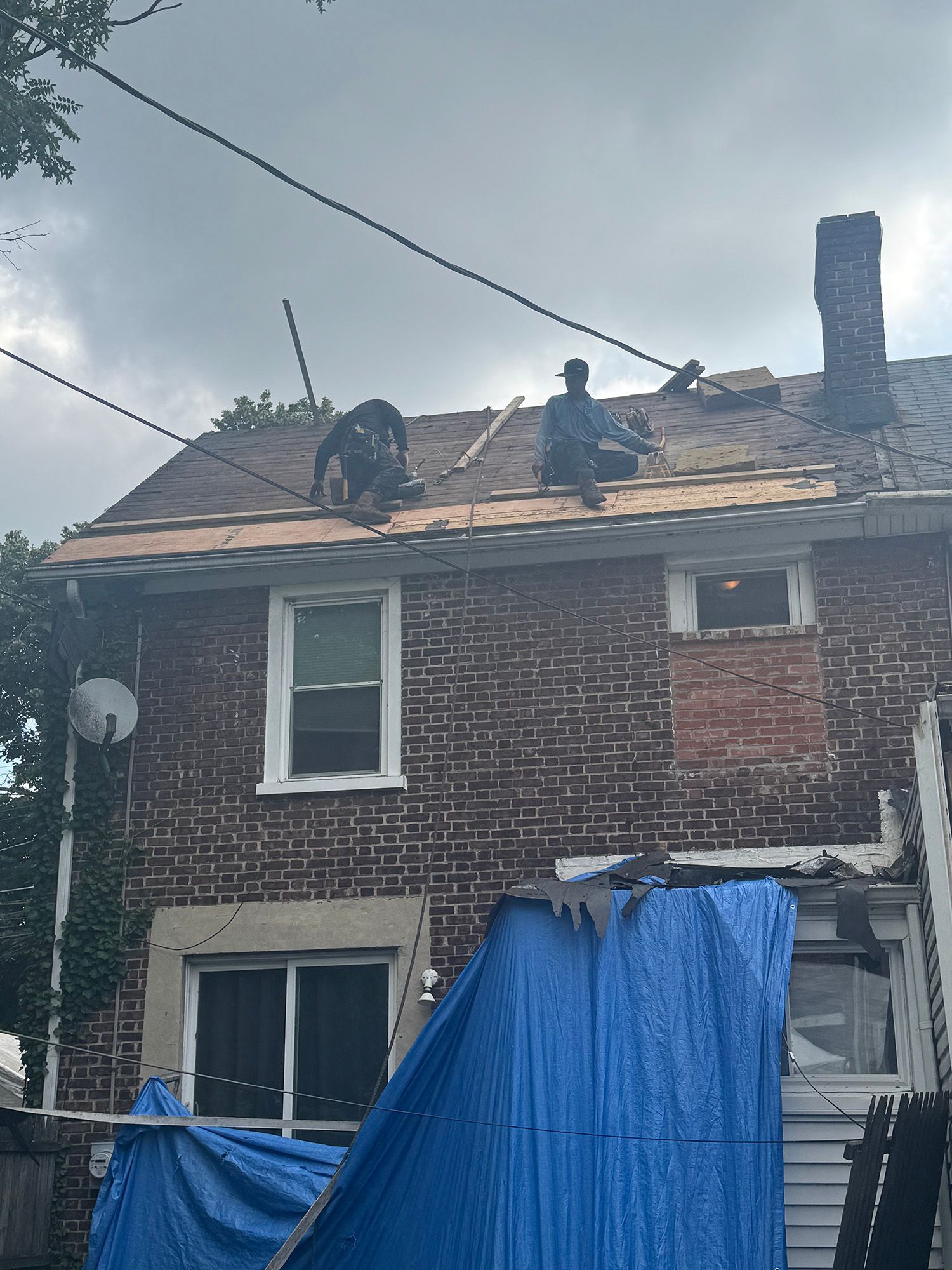 Two roofers repairing a brick house roof under a cloudy sky. A blue tarp covers below.