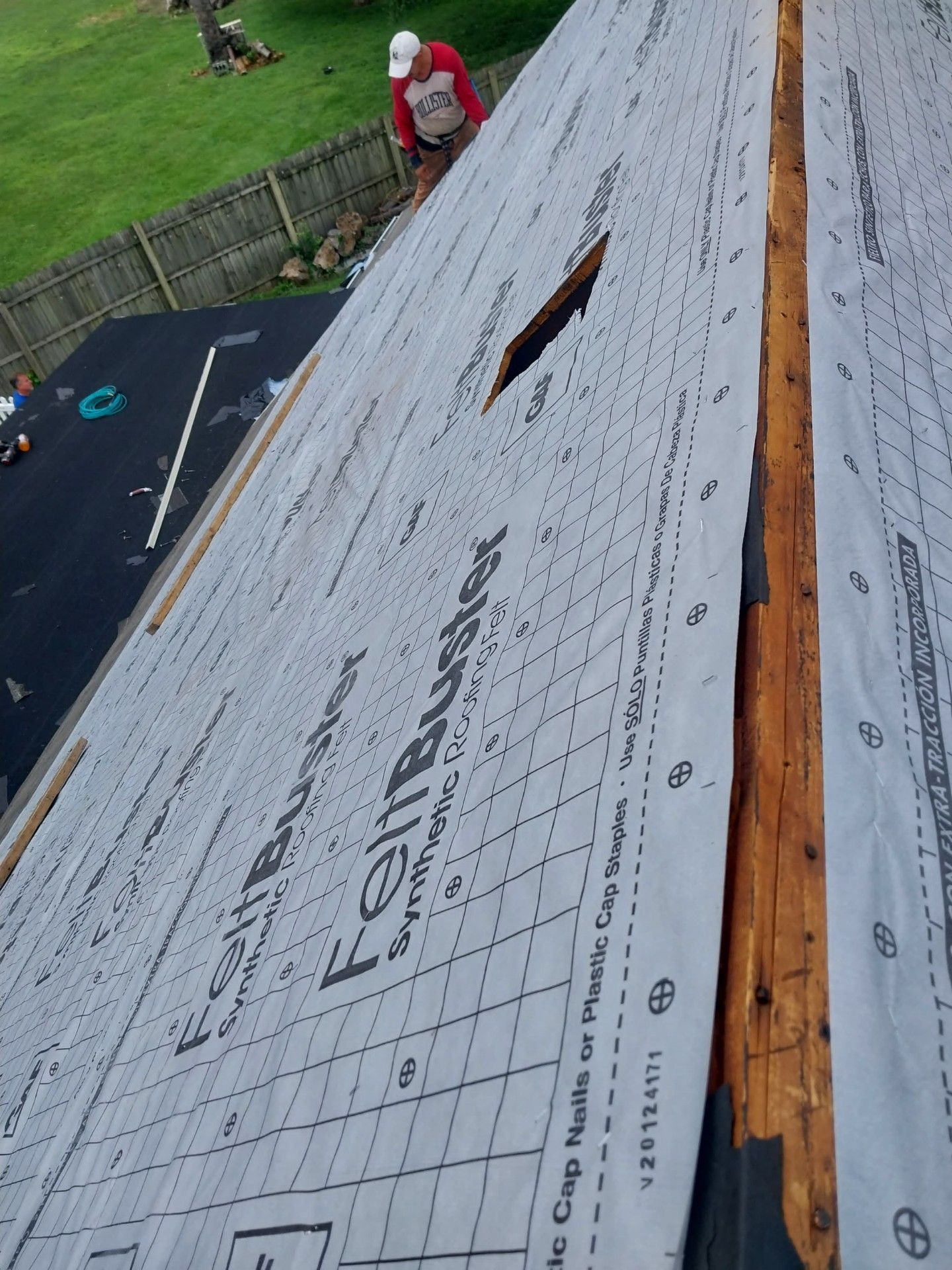 Roofer on a roof, installing underlayment. A hole in the underlayment. Wooden beams and a fence are visible.