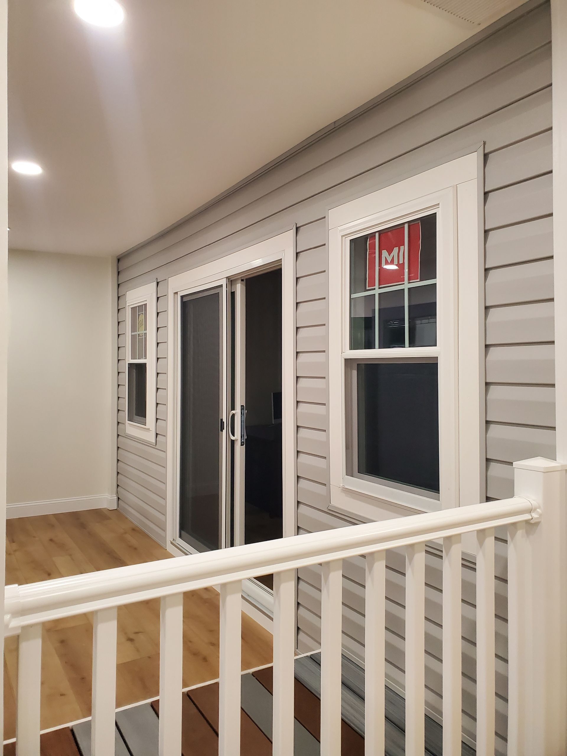 Interior view of a sunroom with gray siding, white trim, and a wooden floor. There's a railing and a sliding glass door.