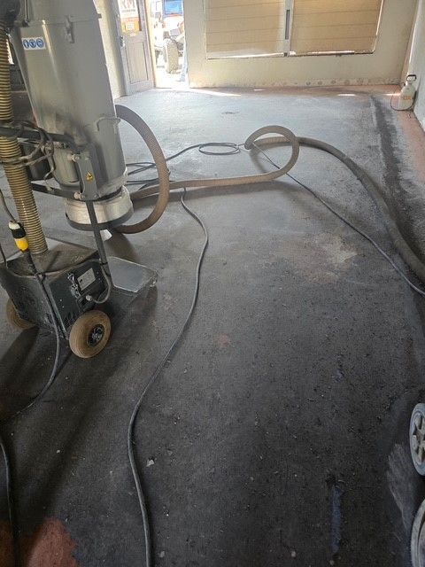 Floor grinding machine in use, dark gray floor, hose and cords, indoor setting.