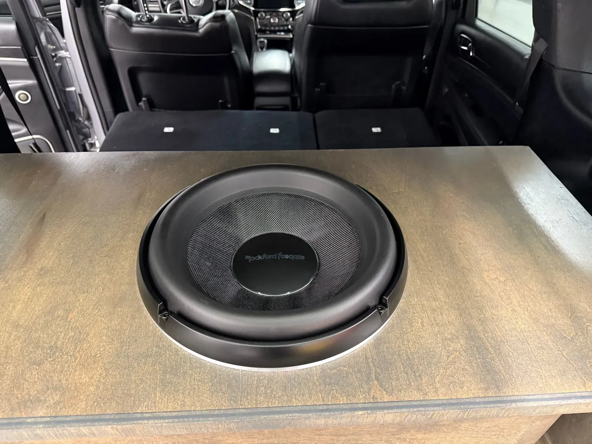A subwoofer installed in a car trunk, mounted in a wooden enclosure. Black speaker, car interior visible.