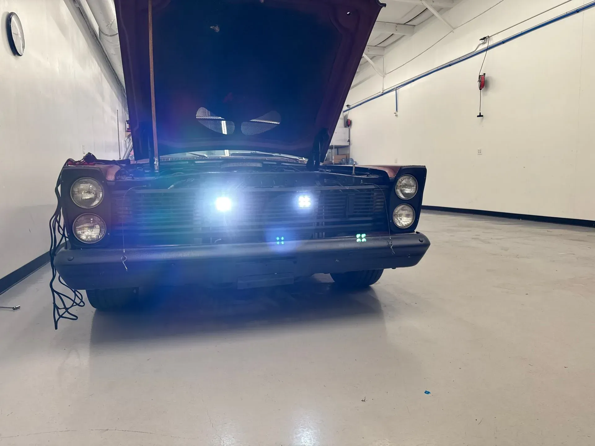 Vintage maroon car with hood up, bright lights on, inside a well-lit garage.