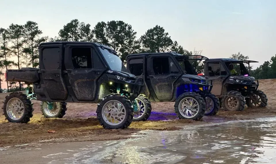 Three lifted black side-by-side ATVs with large chrome wheels parked in muddy terrain.
