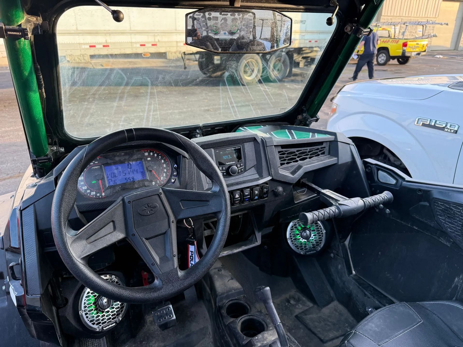 Interior of a green Polaris UTV. Dashboard, steering wheel, and windshield are visible. A truck and person stand outside.