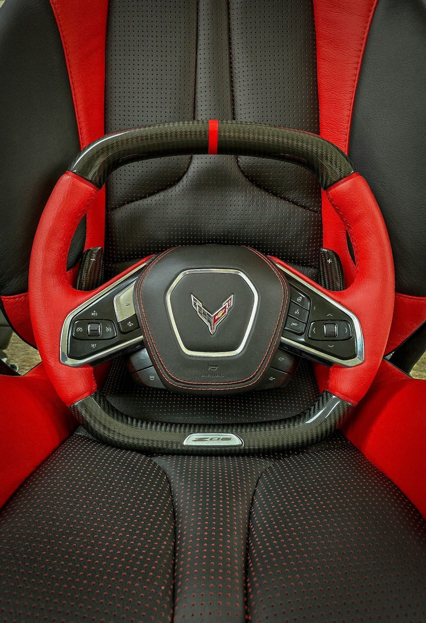 Red and black Corvette steering wheel centered on a black perforated car seat with red accents.