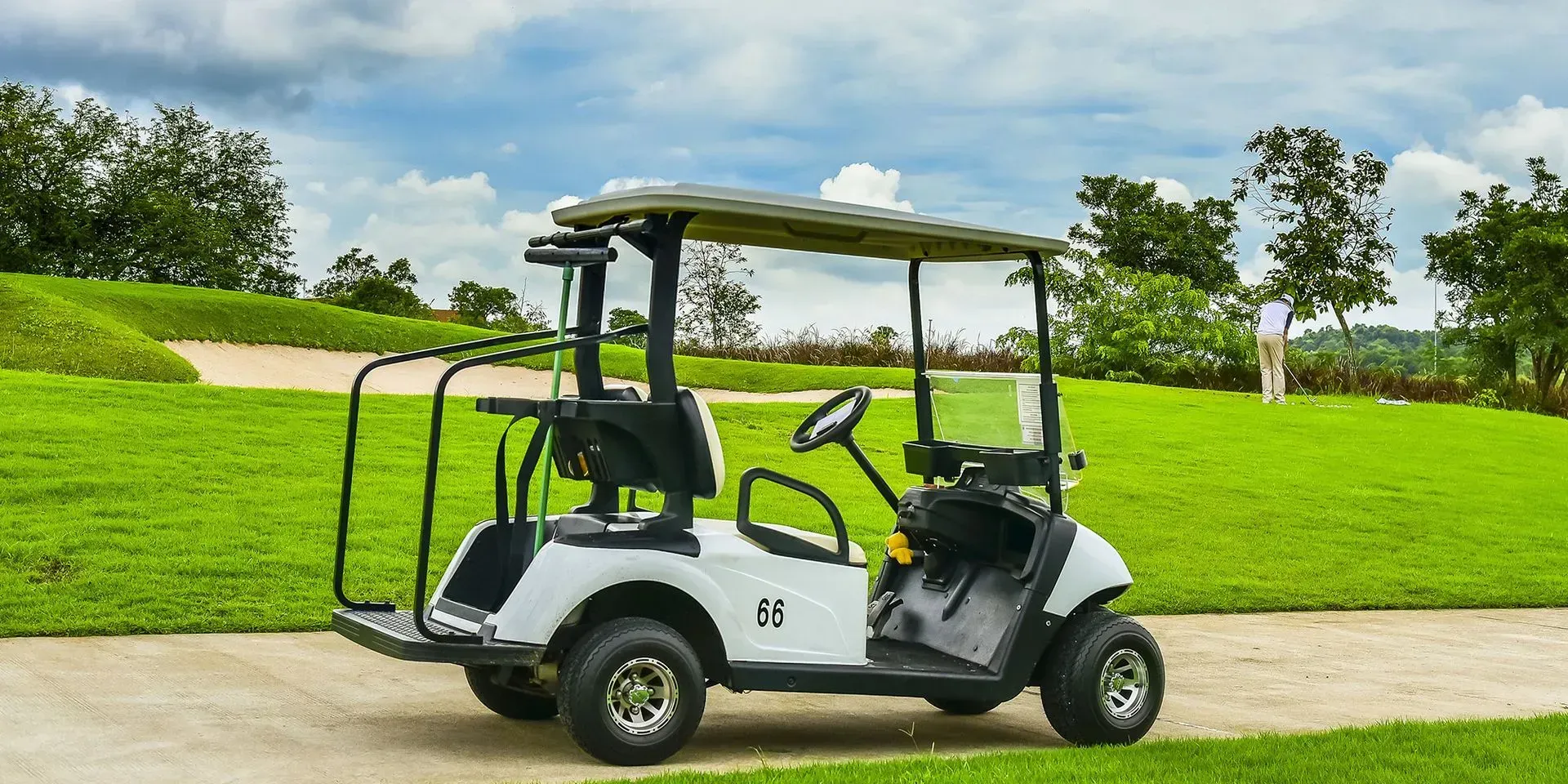 White golf cart on a path, green grass, trees, and a cloudy sky in the background.