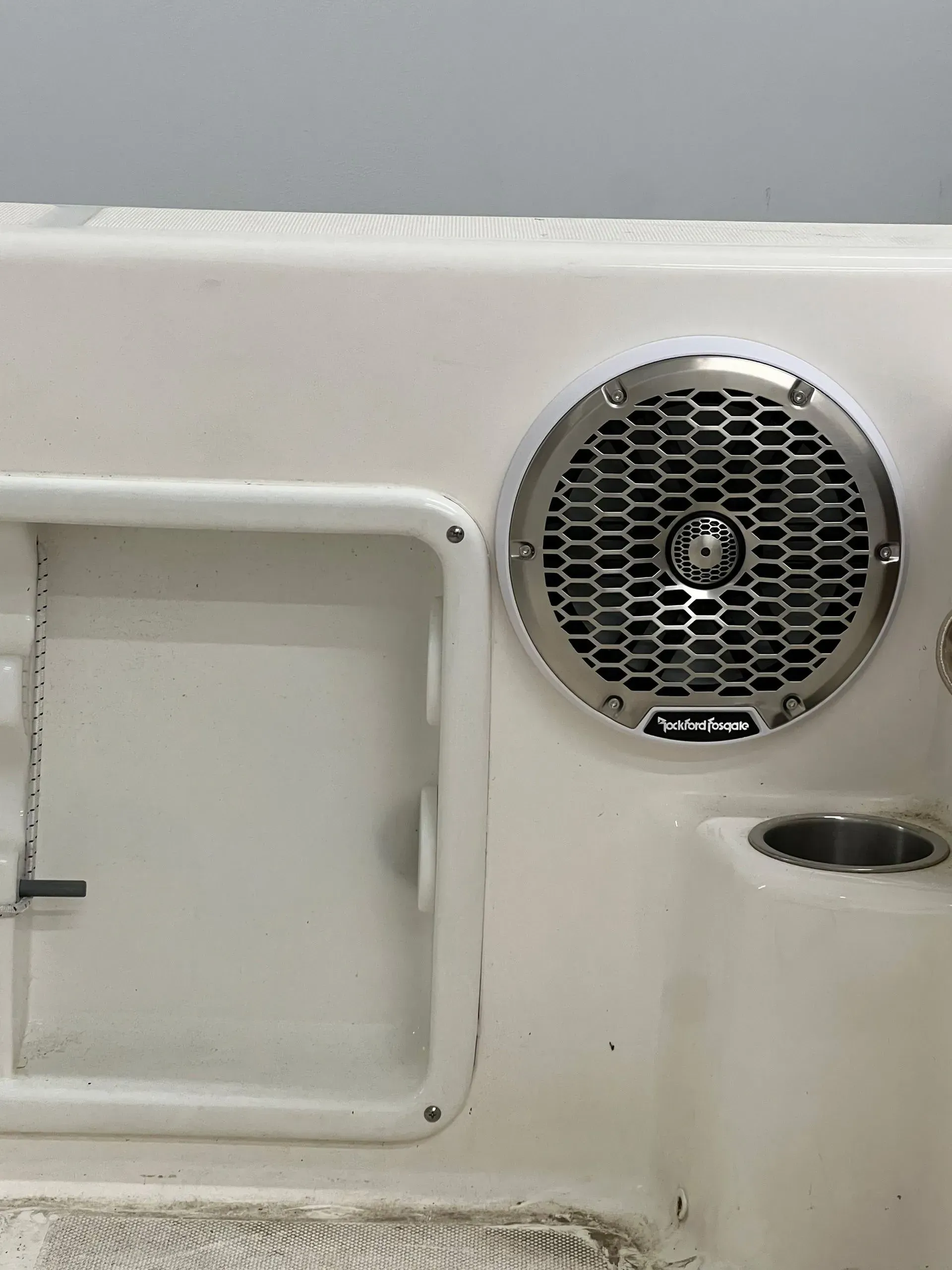 A round speaker with a silver grill on a white boat surface next to a recessed storage area.