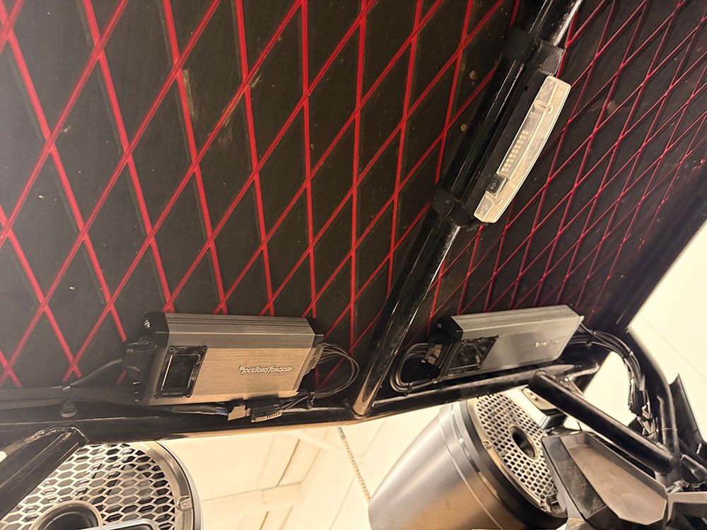 Two silver audio amplifiers mounted on a black roll cage, above speakers. Red diamond stitching on black.