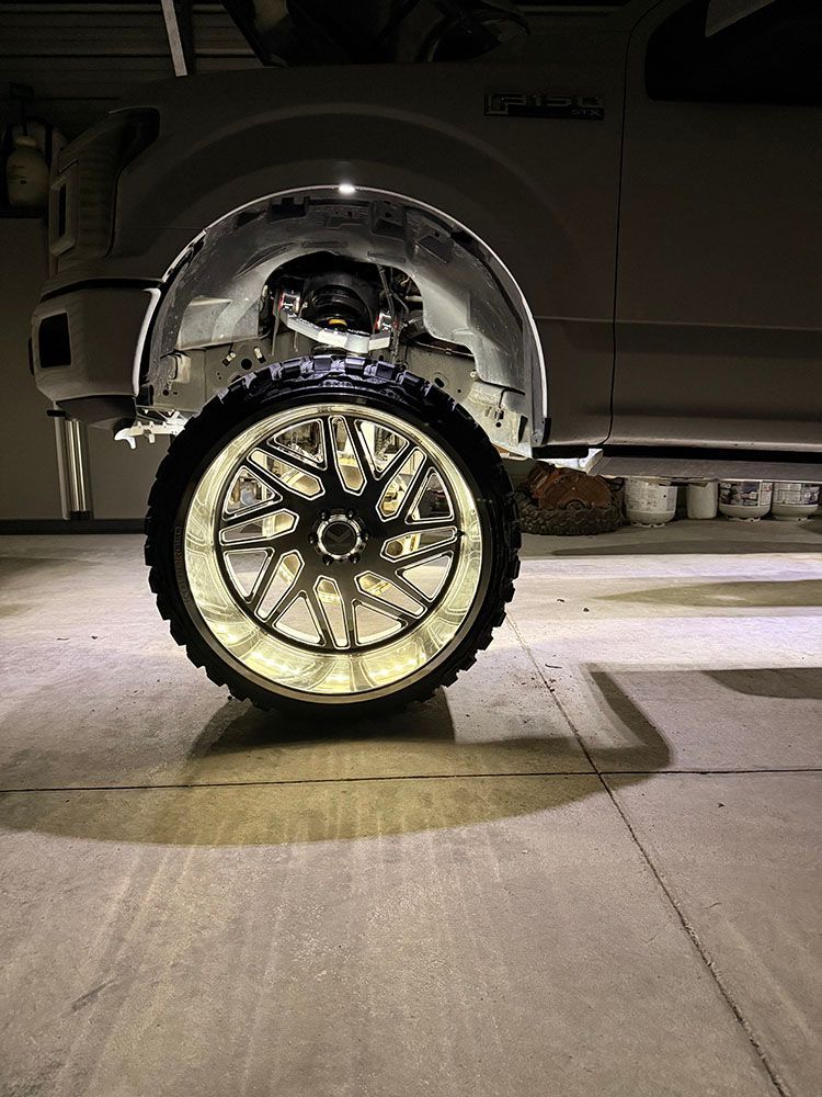 White truck's front wheel with a lighted rim and underglow. Parked indoors, the tire is large with a chrome design.