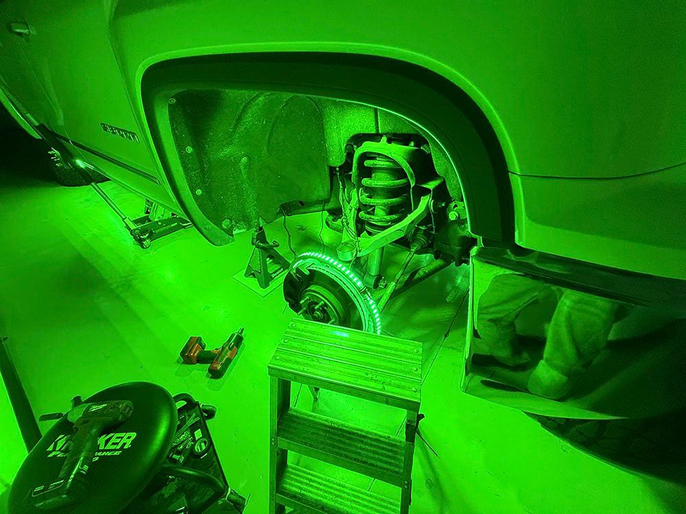 Green-lit view of a car's wheel well, brake assembly exposed, tools, small ladder.