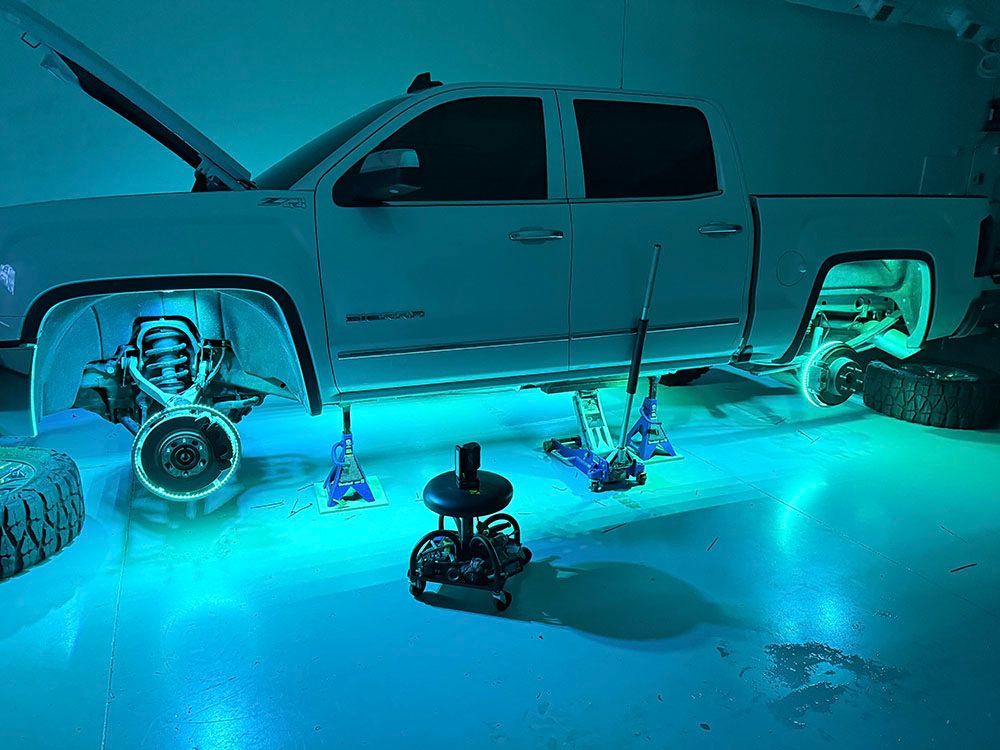 White truck lifted in a garage, illuminated with blue lights. Wheels removed, jack stands supporting it.