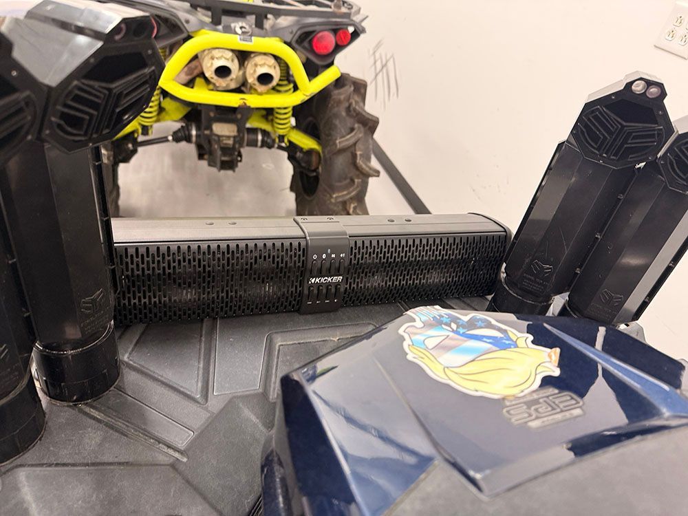 Yellow off-road vehicle over black audio equipment on blue, logo-adorned surface. Black exhaust pipes flank.