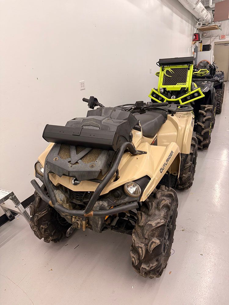 Four tan and black ATVs parked in a row, muddy tires, inside a building.