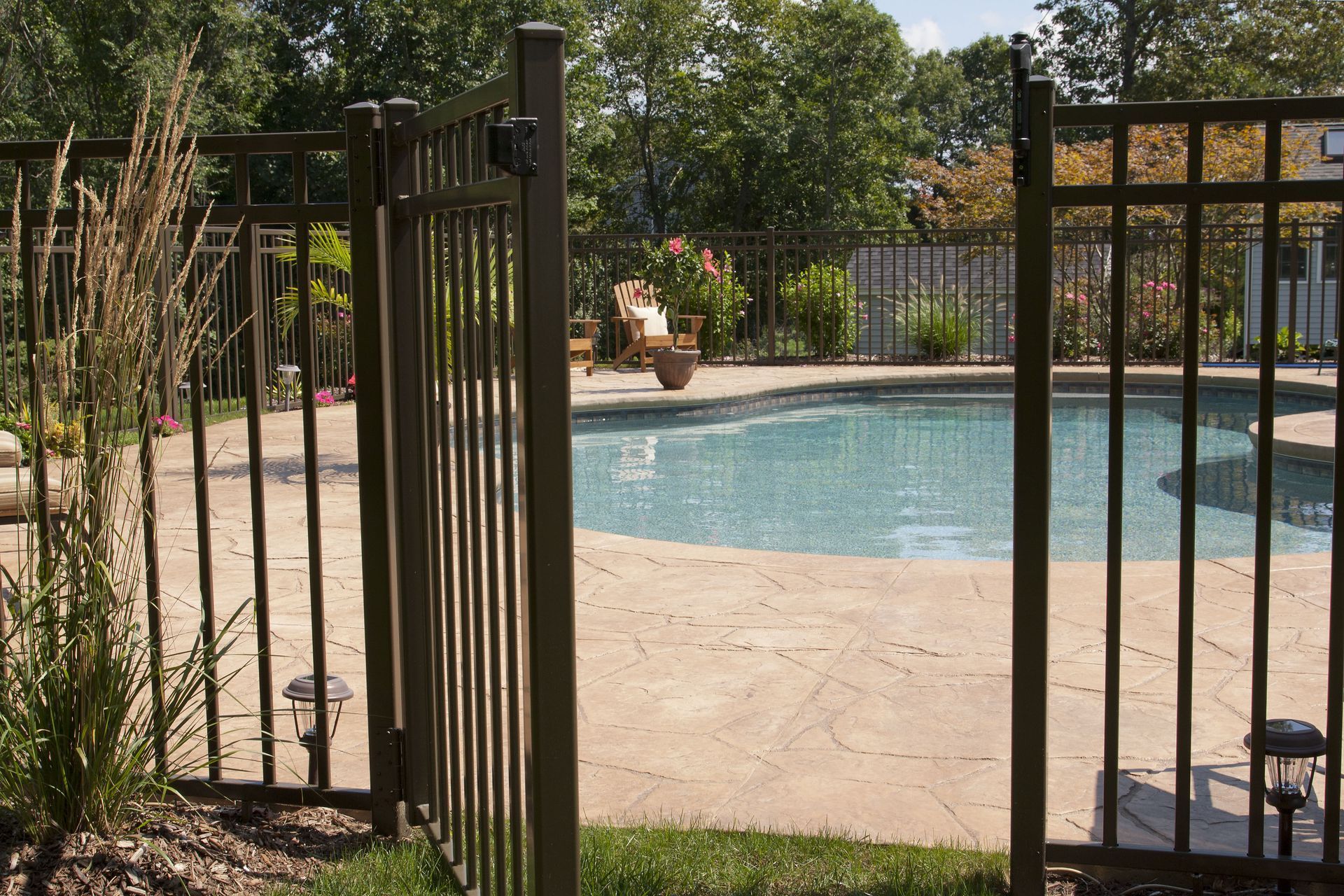 Gated backyard swimming pool with stone patio and surrounding trees on a sunny day