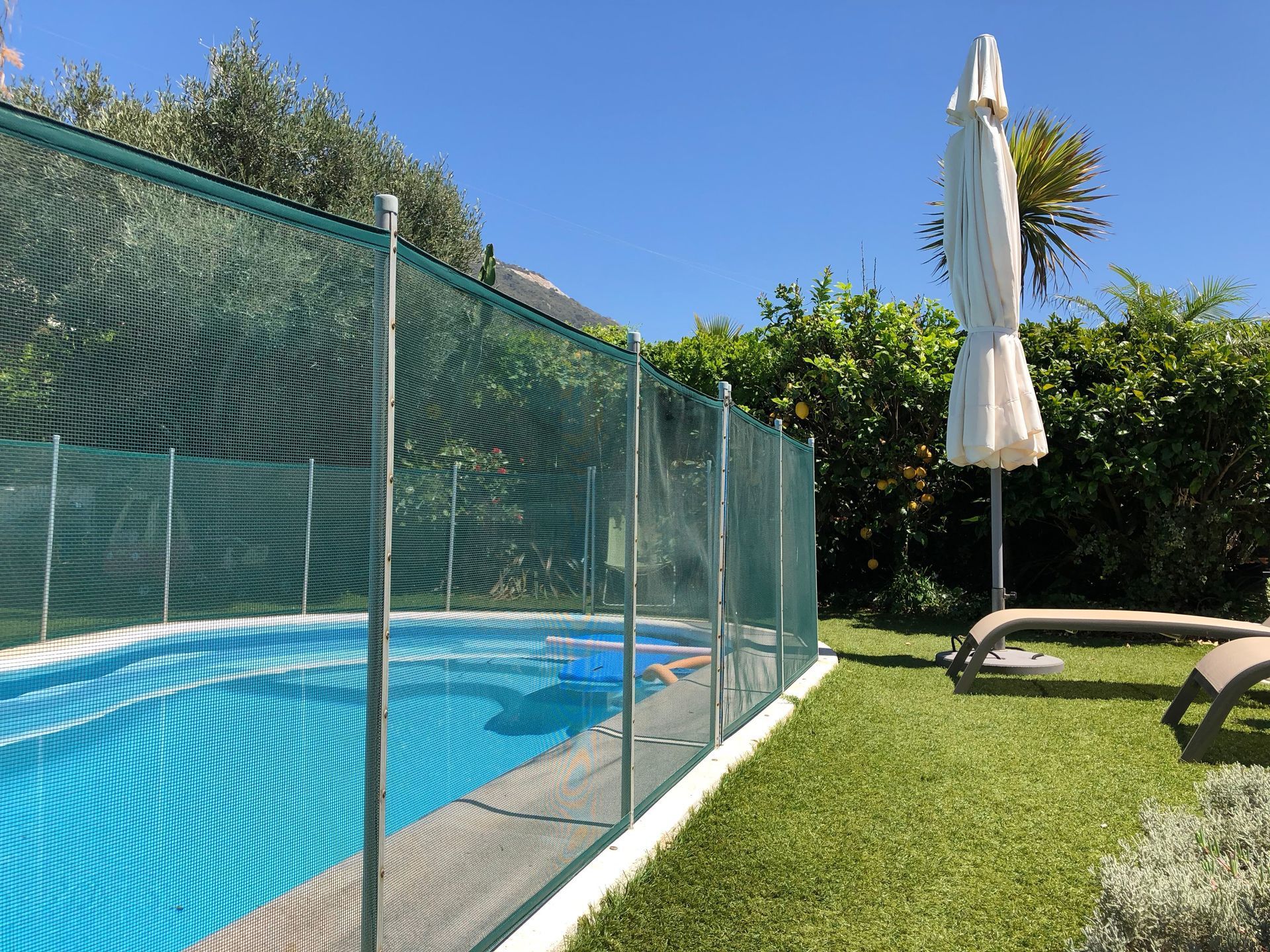 Backyard pool with blue water, green fencing, lawn chairs, and a closed white umbrella
