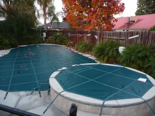Covered backyard pool with teal safety cover, autumn trees, and wooden fence behind it.