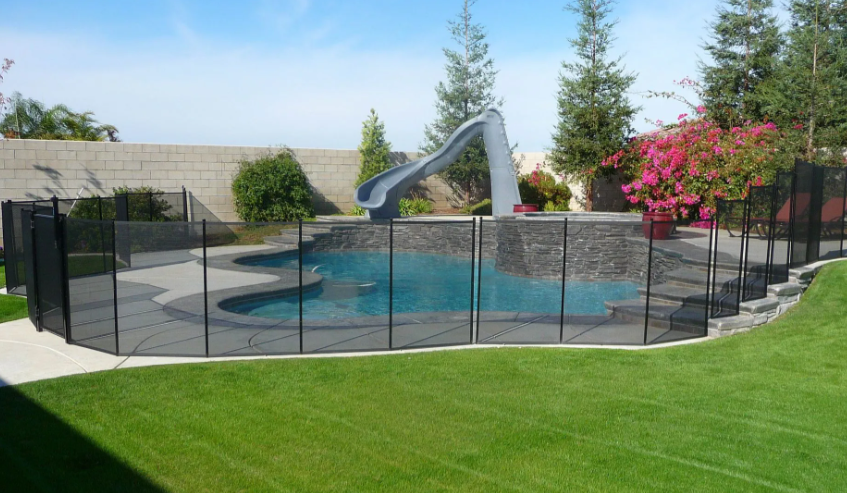 Backyard pool with black safety fence, gray diving slide, and green lawn under a clear sky