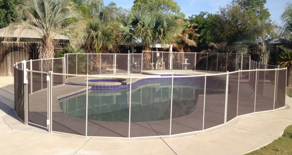 Backyard swimming pool enclosed by a white safety fence, with palm trees and patio around it