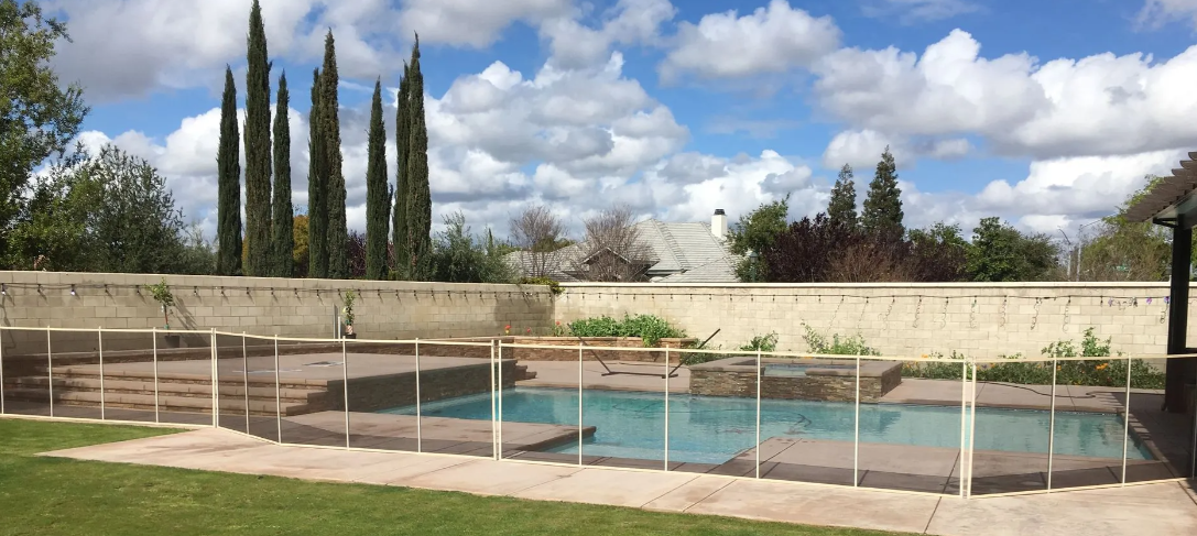 Backyard pool with turquoise water, white fence, stone wall, and tall trees under a cloudy sky